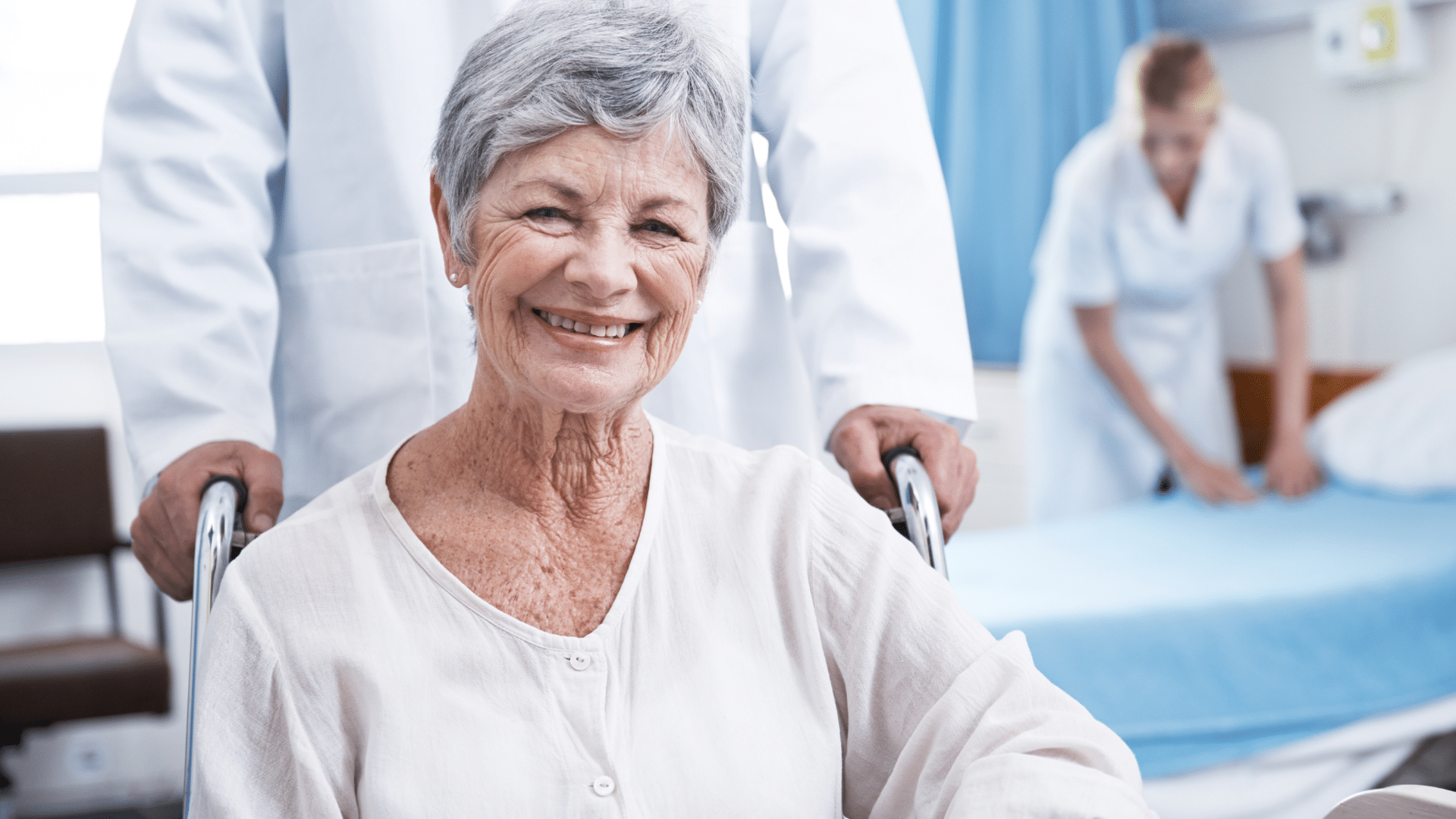 A cheerful elderly woman in a wheelchair accompanied by a nurse, both wearing warm smiles.