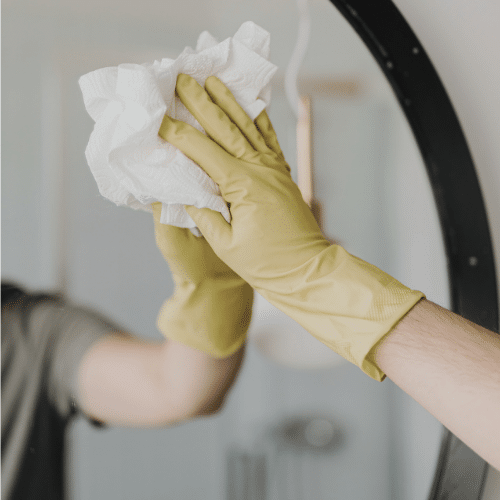 A mirror being cleaned by a person wearing yellow gloves, using a white wipe to remove all the dirt and fingerprints.