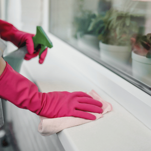 A person wearing pink gloves is cleaning a window sill. They are using a cloth to wipe away dirt and make the window sill spotless.
