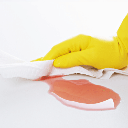 A person with a yellow glove is cleaning a red spill on a white surface using a wipe