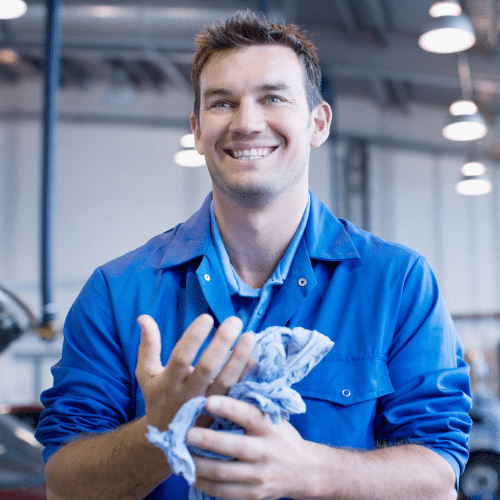 An individual in a workshop environment wearing blue overalls, using a blue cloth to clean his hands.