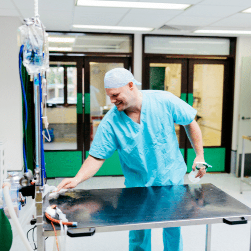 A medical professional in scrubs, cleaning a metal table in a medical setting.