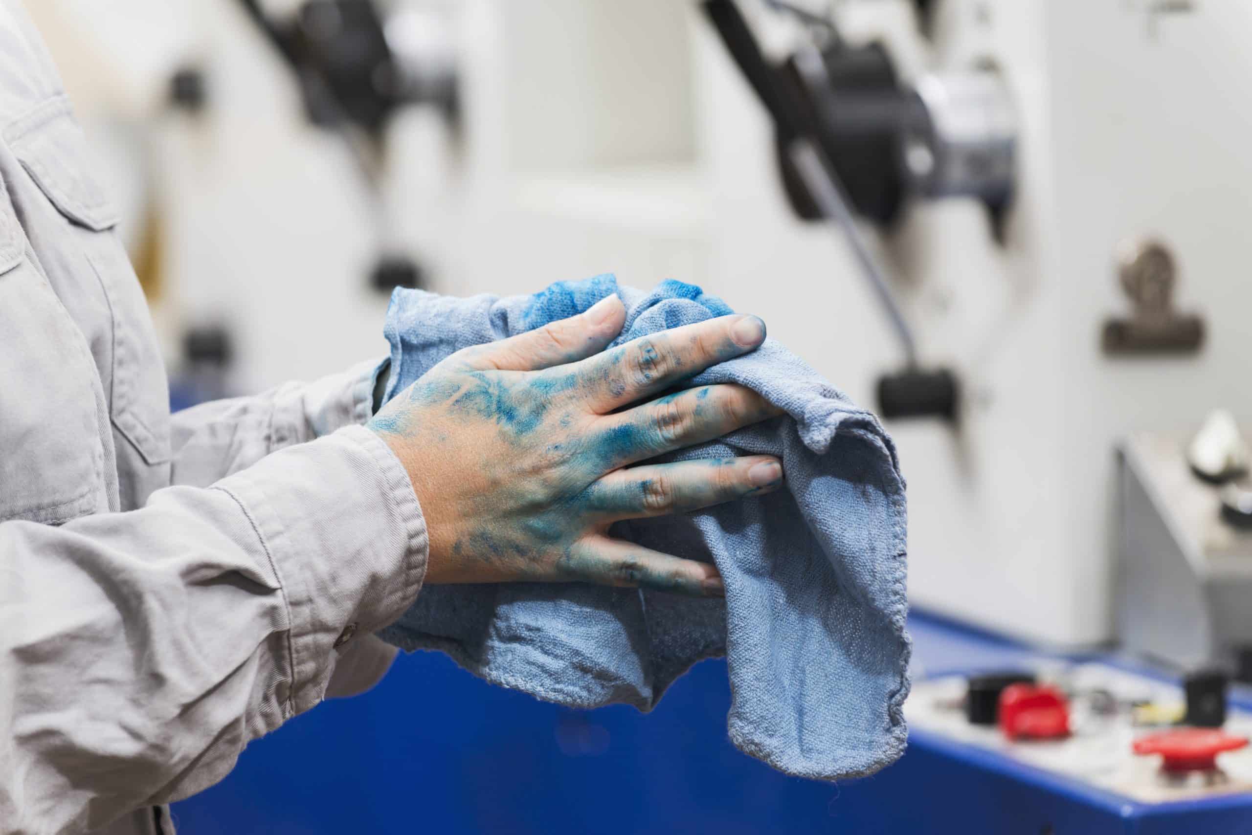 Hands of a female worker in a printing plant, splattered with blue ink. She is wiping her hands with a rag. Machinery is out of focus on background.