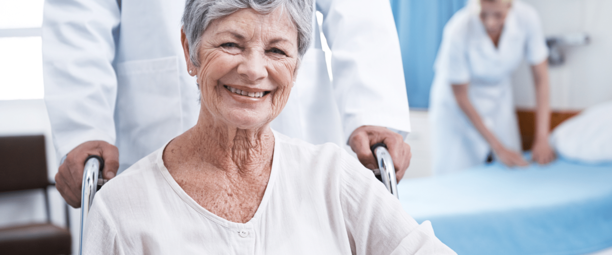 A cheerful elderly woman in a wheelchair accompanied by a nurse, both wearing warm smiles.